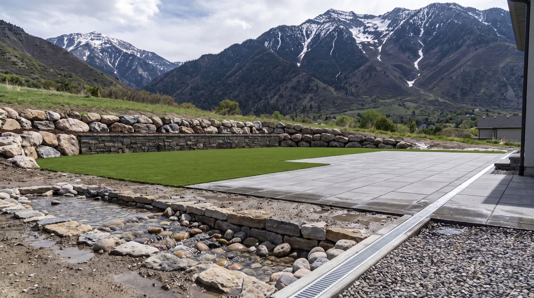 A backyard on the bench below a Wasatch canyon mouth in late spring, turf lawn and paver terrace above a stone-lined swale carrying snowmelt, snow still streaking the peaks behind